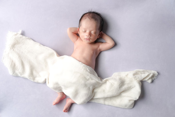 一张专业的新生儿照片捕捉到一位半个月大的日本宝宝安静地睡在放松的姿势中，使用柔软的包巾作为毯子，背景是淡紫色，配以柔和梦幻的灯光。图片下载
