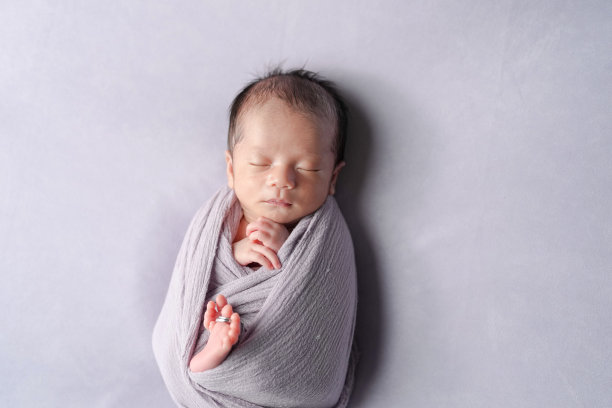 一张专业的新生儿照片展示了一名半个月大的日本婴儿安静地睡着，裹在柔软的布料中。在柔和的浅紫色背景下，婚戒静静地放在婴儿的小脚趾上。图片下载