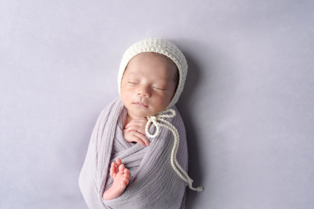 一张专业的新生儿照片，展示了一名半个月大的日本宝宝安静地睡着，裹在柔软的包裹中。宝宝戴着一顶简单的白色针织帽，靠在淡紫色的背景上。图片下载