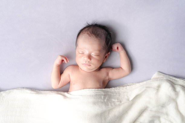 一张专业的新生儿照片捕捉到一位半个月大的日本宝宝安静地睡在放松的姿势中，使用柔软的包巾作为毯子，背景是淡紫色，配以柔和梦幻的灯光。图片下载