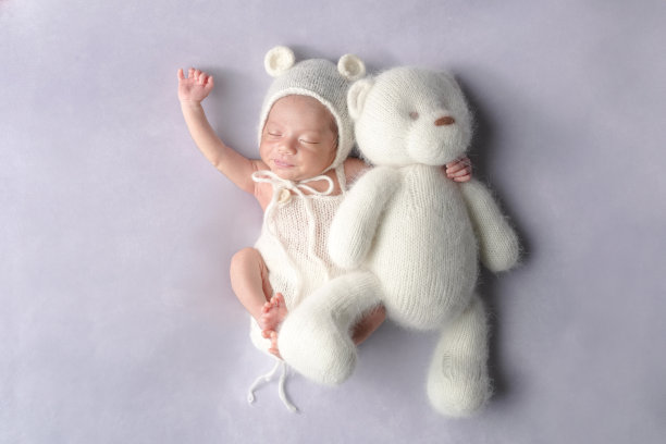一张专业拍摄的新生儿照片，展示了一名半个月大的日本宝宝穿着熊装，安静地躺在一个大型泰迪熊旁边，背景是柔和的浅紫色，专为新生儿摄影而设计。图片下载