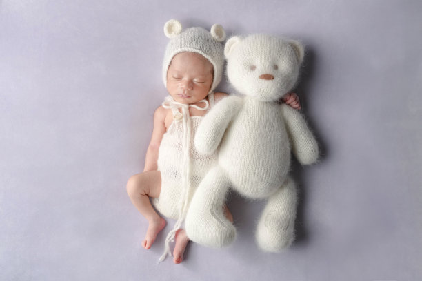 一张专业拍摄的新生儿照片，展示了一名半个月大的日本宝宝穿着熊装，安静地在柔和的浅紫色背景上与一只大泰迪熊一起入睡，背景专为新生儿摄影而设。图片下载