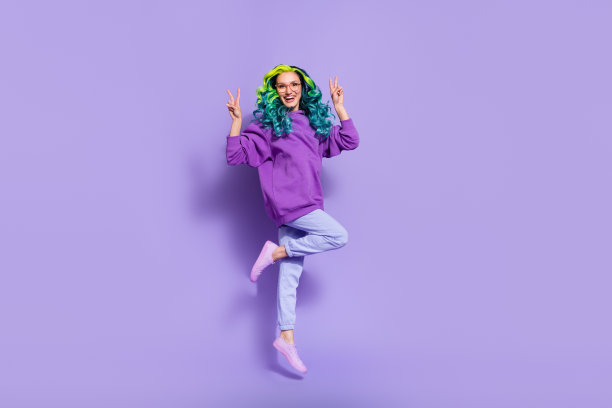 充满活力的年轻女孩在紫色背景上跳起，做出V字手势的全身照片图片下载