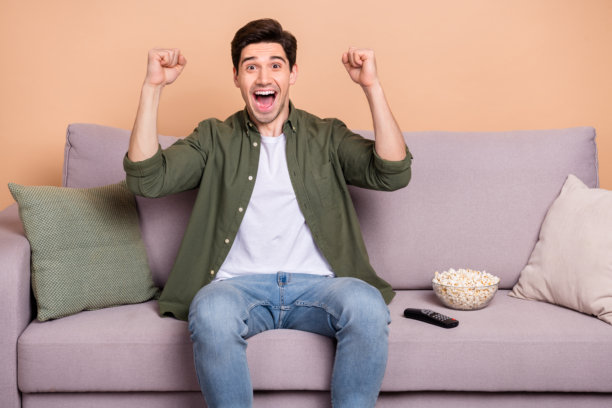 兴奋的男孩坐在沙发上，握拳观看世界杯比赛，享受国家队的胜利，背景为米色。图片下载
