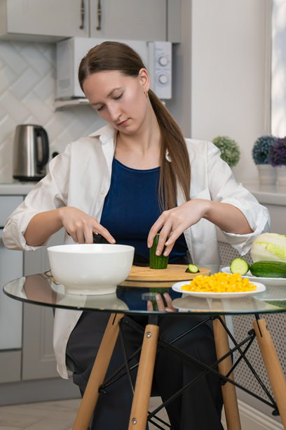 年轻女性在现代厨房中准备健康沙拉，正在木砧板上切新鲜黄瓜，周围还有玉米和北京白菜等其他食材。竖版照片图片下载