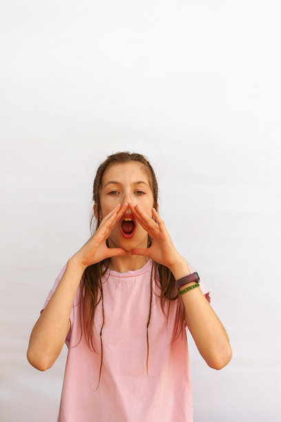 一个十几岁的女孩像在对着扩音器尖叫，双手捂住脸，嘴巴张开。白色背景上的竖版照片。图片下载