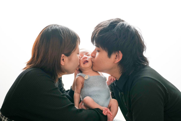 白色背景下的两周大婴儿照片，婴儿穿着蓝色连体衣，被一对年轻的日本夫妇温柔地抱着。两人从两侧亲吻婴儿。温馨而专业的构图。图片下载