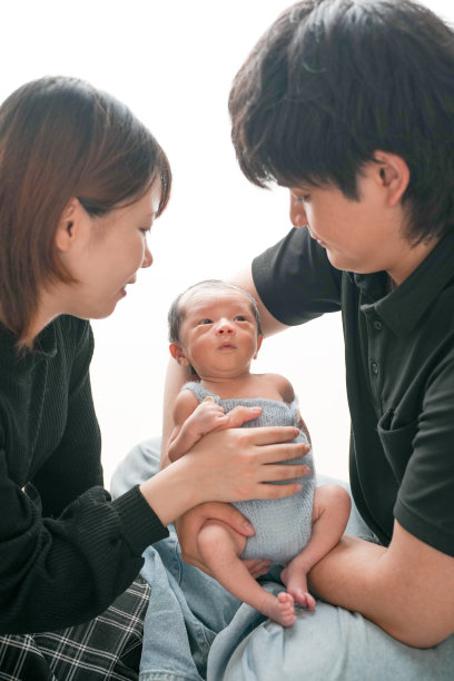 白色背景上的新生儿照片，展示了一名两周大的婴儿穿着蓝色连体衣，温柔地被一对二十多岁的年轻日本父母抱着。干净而温馨的专业构图。图片下载