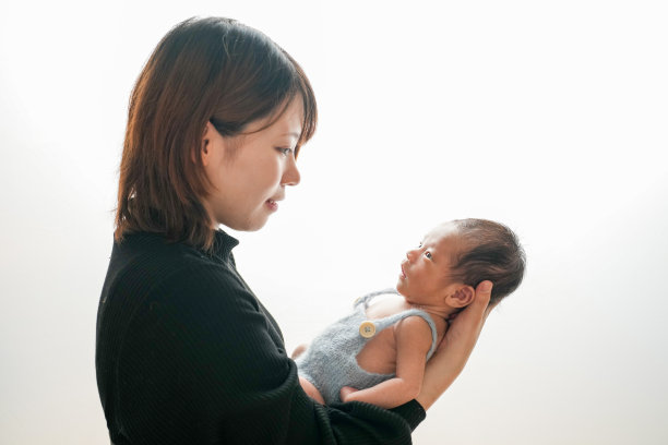 白色背景上的新生儿照片，展示了一名两周大的婴儿穿着蓝色连体衣，温柔地被一位二十多岁的年轻日本母亲抱着。干净、简约的构图，营造出温馨的氛围。图片下载