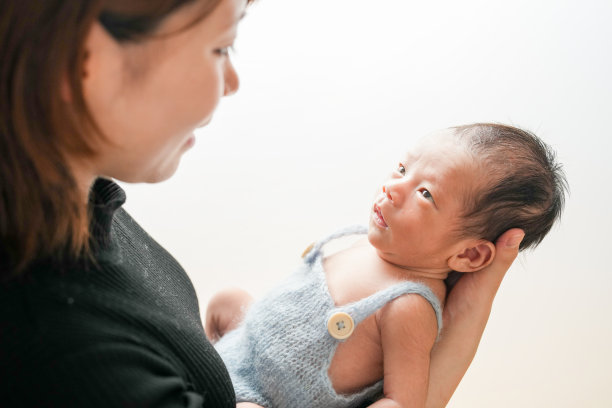 白色背景上的新生儿照片，展示了一名两周大的婴儿穿着蓝色连体衣，温柔地被一位二十多岁的年轻日本母亲抱着。干净、简约的构图，营造出温馨的氛围。图片下载