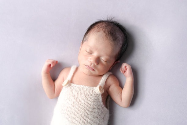 一张专业拍摄的新生儿照片，展示了一名半个月大的日本宝宝穿着白色连体衣，安静地睡在柔和的浅紫色背景上，捕捉了新生命的宁静与细腻之美。图片下载