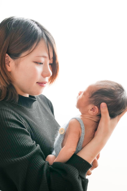 白色背景上的新生儿照片，展示了一名两周大的婴儿穿着蓝色连体衣，温柔地被一位二十多岁的年轻日本母亲抱着。干净、简约的构图，营造出温馨的氛围。图片下载