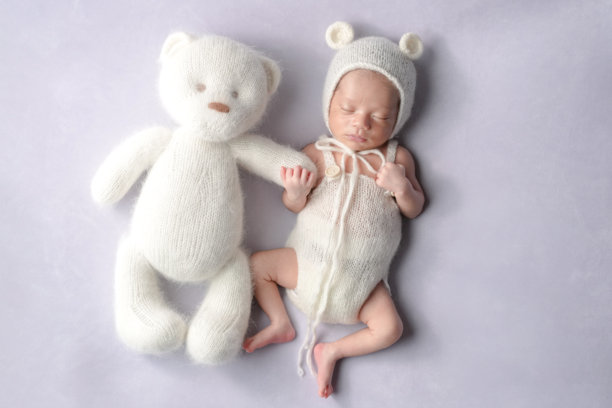 一张专业拍摄的新生儿照片，展示了一名半个月大的日本宝宝穿着熊装，安静地在柔和的浅紫色背景上与一只大泰迪熊一起入睡，这个背景专为新生儿摄影而设计。图片下载