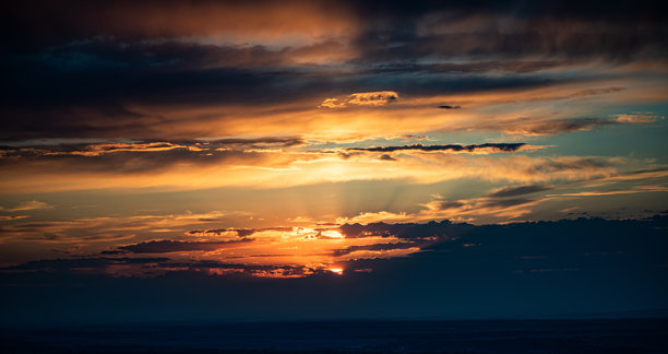 这张照片捕捉了日落时分，明亮的橙色和金色阳光穿透层叠的云层。地平线昏暗，天空充满戏剧性的光影和丰富的色彩。图片下载