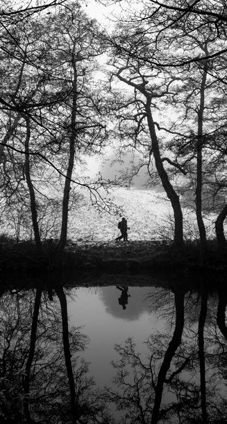 黑白冬季照片，展示两个人在英国德比郡德文特河岸边走过，树木之间，背景是美丽的倒影和一片雪地。图片下载