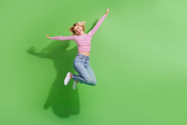 一位无忧无虑、光彩照人的女士穿着条纹上衣，在空旷的绿色背景下高高跳起的全身照片图片下载