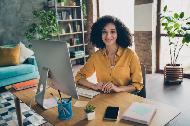 一位迷人的自由职业女性穿着衬衫，微笑着在室内工作站使用现代设备发信息的照片图片下载
