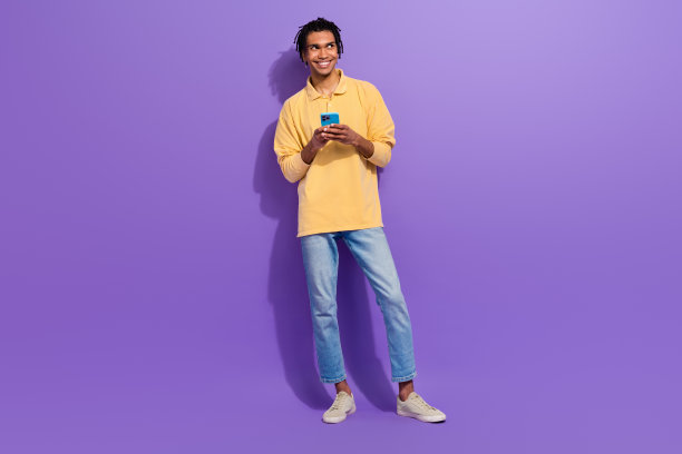 穿着橙色衬衫和牛仔裤的年轻博主，手持智能手机，梦幻般地站在紫色背景前，展示全身照片，正在进行在线购物。图片下载