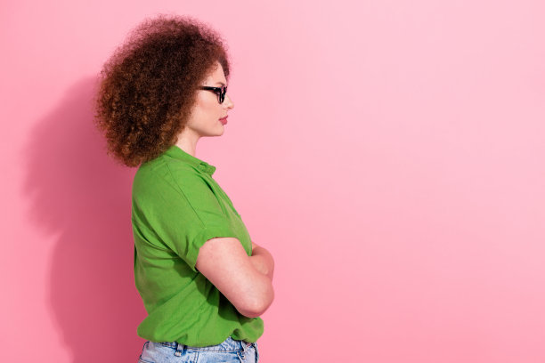 年轻女士的照片肖像，双手交叉，表情严肃，身穿时尚的绿色服装，背景为粉色，留有空白空间。图片下载