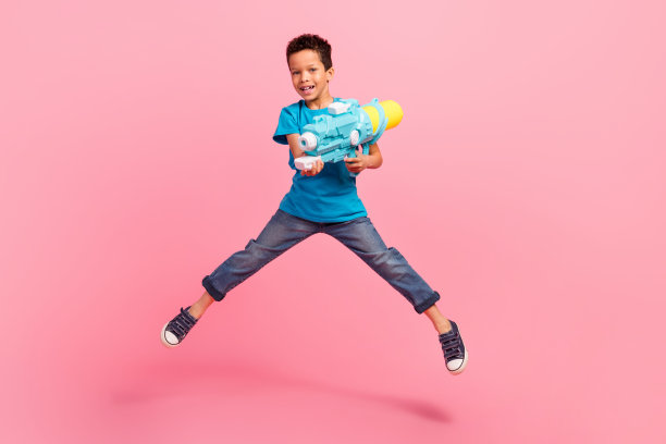 全身照片，快乐的时尚小男孩穿着蓝色T恤，手持水枪高高跳起，背景为粉色。图片下载