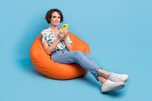 可爱的积极女性穿着印花衬衫，使用苹果或三星手机发短信，背景为空白的蓝色。图片下载