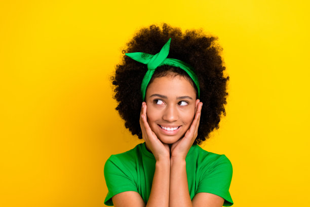 梦幻而沉思的女士，穿着绿色T恤，戴着发带，双臂托着脸颊，凝视着空旷的空间，背景为黄色。图片下载