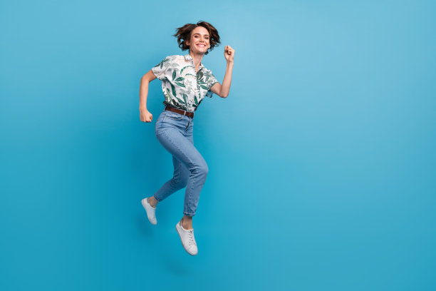 兴奋的酷女孩穿着印花衬衫在空旷的蓝色背景下高高跳起，快速奔跑的全身照片图片下载