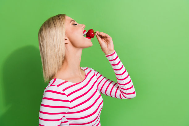 侧面照片，漂亮的年轻女士，金色短发，咬了一小口草莓，夏季甜美成熟的水果，背景为绿色。图片下载