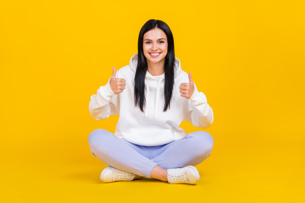 全身照片的女士坐着，双腿交叉，竖起大拇指，手势暗示选择，背景为明亮的黄色。图片下载