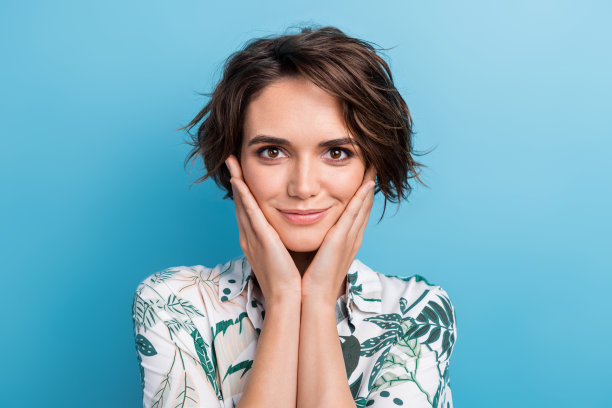 可爱的年轻女士的照片肖像，双手托着脸颊，欣赏着穿着时尚印花叶子图案的服装，背景为蓝色。图片下载