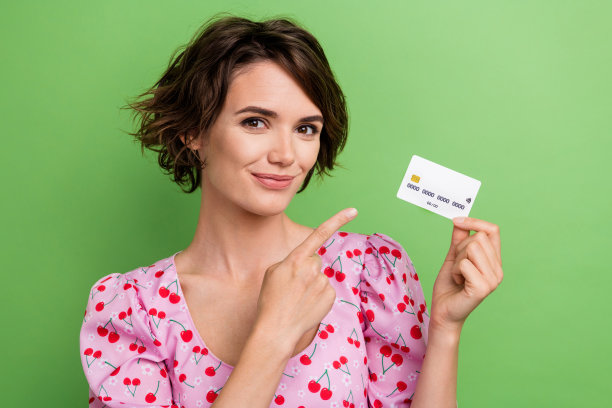 可爱的年轻女士在绿色背景下穿着时尚的粉色樱桃印花服装，手指指向信用卡的照片肖像。图片下载
