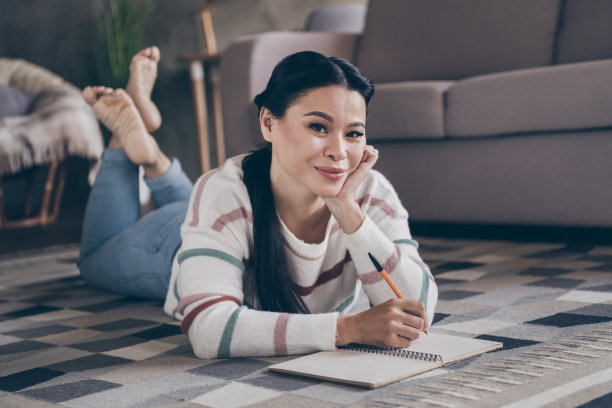 年轻迷人的女士全身侧面照片，躺在室内自我隔离公寓的地板上，正在写作。图片下载