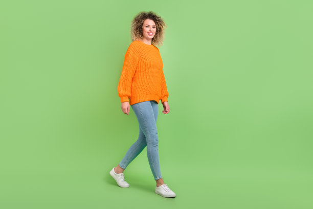 穿着橙色毛衣、牛仔裤和鞋子的金色卷发女士的全身照片，背景为绿色。图片下载