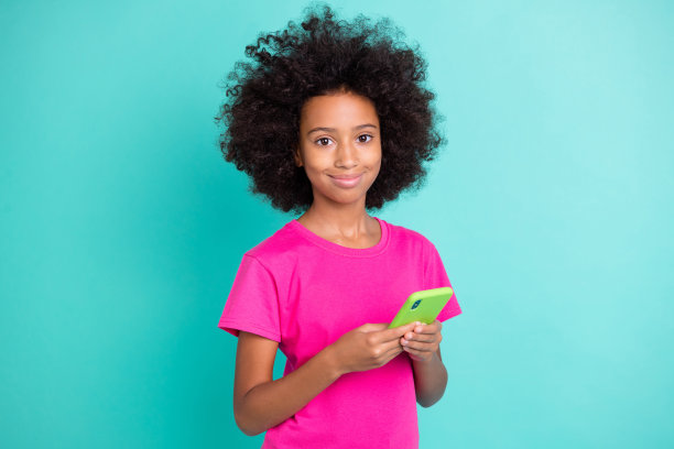 积极的黑肤女孩双手握着手机的照片肖像，背景为鲜艳的青色。图片下载
