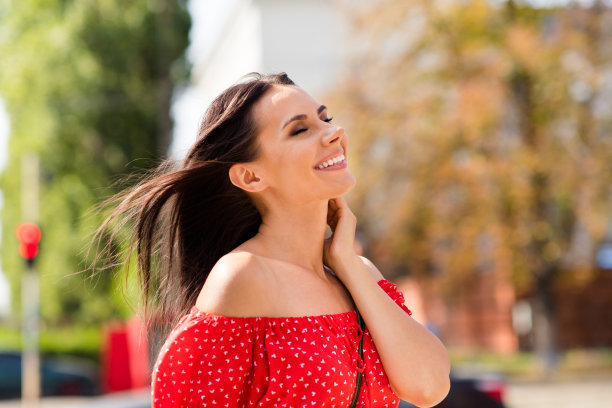 侧面肖像照片，快乐的女性微笑大笑，轻触脖子，在风大的天气中闲逛于城市街道。图片下载