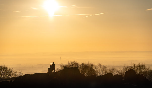 一个男人将他的狗放在基座上，享受金色日出美景的剪影照片，背景是朦胧的雾气和风力发电机。图片下载