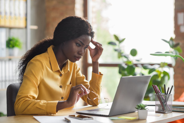 现代办公室内，一位非裔美国女性在使用笔记本电脑时触摸头部的照片肖像。图片下载