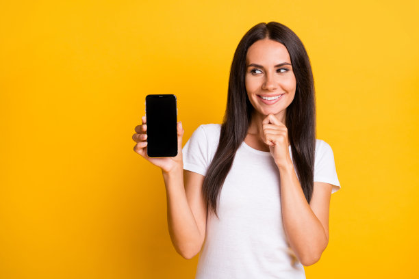 年轻女孩的照片肖像，展示手机触摸屏，手指触碰下巴，背景为明亮的黄色，留有空白空间。图片下载