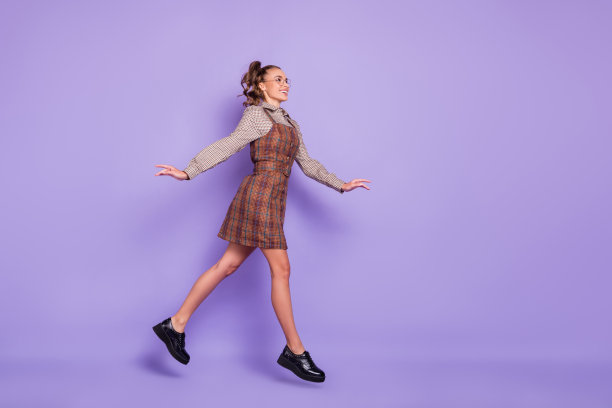 乐观的棕发女孩穿着眼镜、棕色连衣裙和鞋子，完整的侧面照片，背景为淡紫色。图片下载