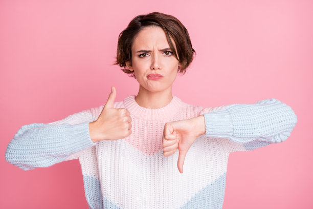 困惑女孩的照片肖像，竖起大拇指和拇指向下，背景为柔和的粉色。图片下载