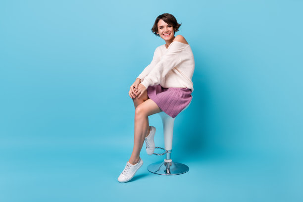 全身肖像照片，快乐的女孩坐在酒吧凳上，背景为柔和的蓝色。图片下载