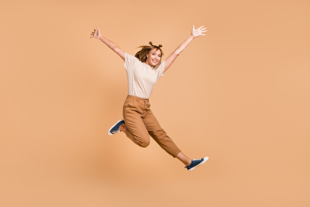 女孩穿着T恤、裤子和运动鞋在米色背景上跳跃，尽情享受乐趣的全身照片图片下载