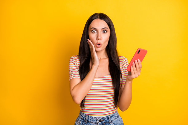 女性博主惊讶地捂着脸颊，手持手机的照片肖像，背景为明亮的黄色。图片下载