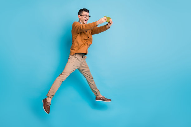 年轻男子全身侧面照片，开心兴奋地跳跃玩手机游戏，背景为蓝色。图片下载