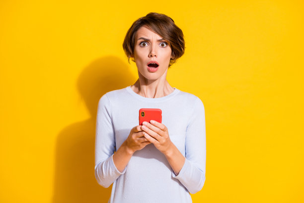惊讶的女孩双手握着手机的照片肖像，背景为明亮的黄色。图片下载