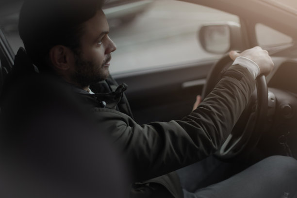 年轻男子侧面驾驶汽车，穿着深色夹克，专注于道路。照片捕捉了一个平静而专注的驾驶时刻，柔和的自然光线洒在车内。图片下载