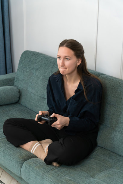 专注的年轻女性舒适地坐在沙发上，手持无线控制器玩视频游戏，享受在家中的闲暇时光，完全沉浸在她最喜欢的爱好中。竖版照片图片下载