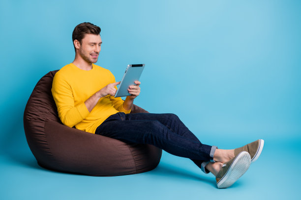 坐在大软椅上的积极人士全身侧面照片，使用平板电脑，背景为蓝色。图片下载