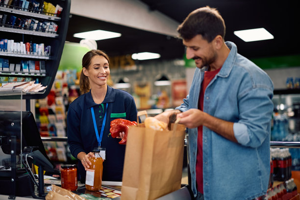 快乐的超市收银员在结账时帮助顾客将杂货装入纸袋。图片下载