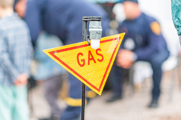 一张坦率的图片，显示一个黄色的气体危险标志，上面有红色字母，遮挡了一个涉及多名穿制服的人员（可能是执法或紧急服务人员）在一个地点的紧急响应场景。图片下载